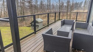 Una terraza de madera con muebles de mimbre y barandilla de vidrio que da a un área verde con árboles.