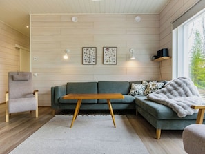 Un salon moderne avec un canapé d’angle gris, une table basse en bois et un fauteuil gris. Deux lampes murales et des reproductions botaniques encadrées sont accrochées au mur au-dessus du canapé.