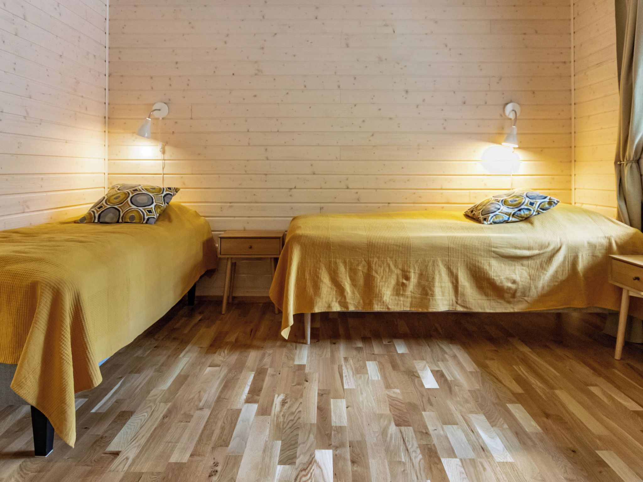A room with two single beds, wooden flooring, and wall-mounted lamps.