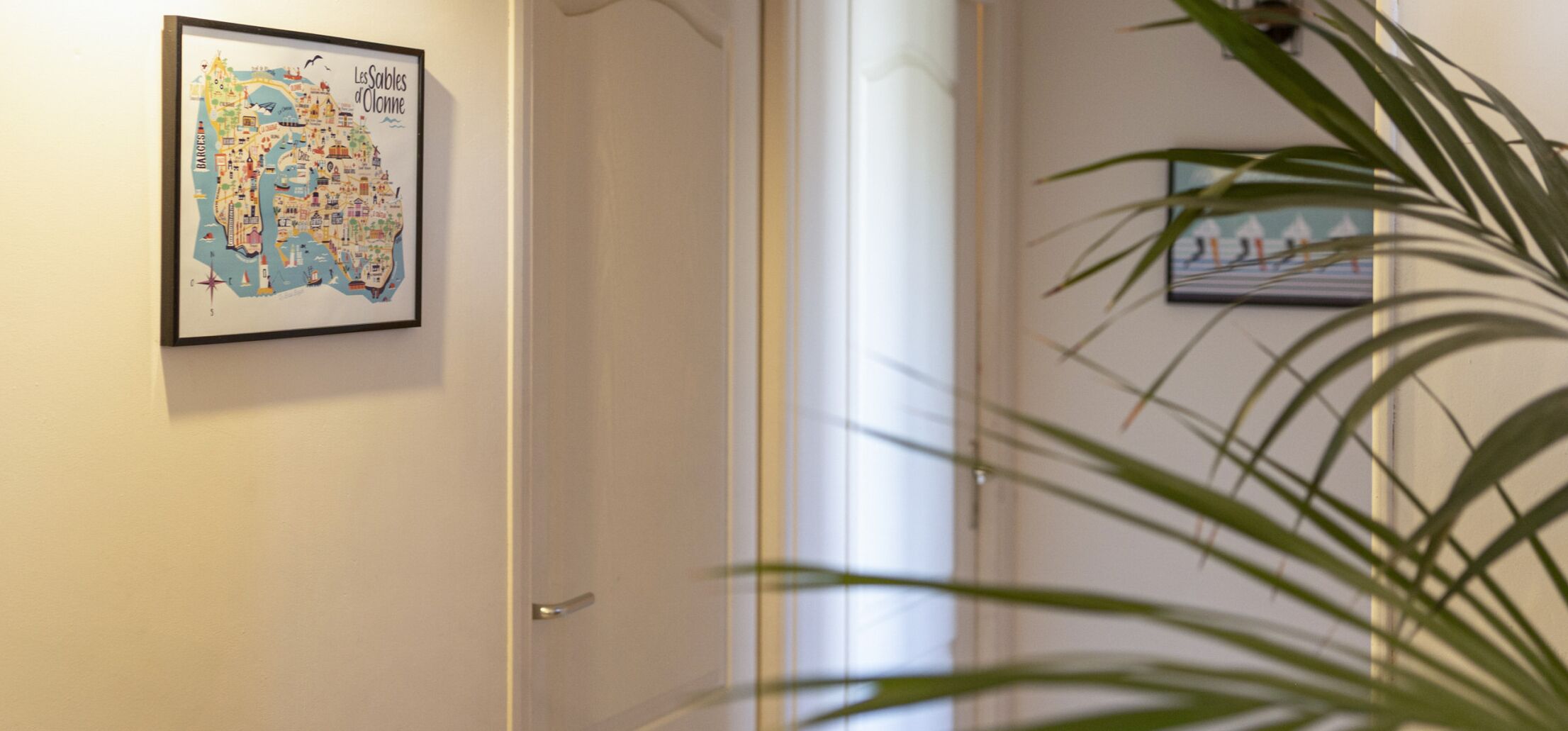 Une carte encadrée de Les Sables-d’Olonne est accrochée au mur, avec une porte et une plante au premier plan.