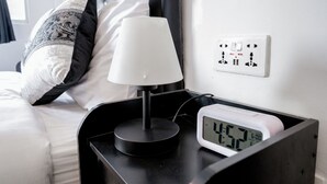 Un letto rifatto con cura, con un poggiatesta nero, un cuscino bianco, un comodino nero e una lampada bianca, e una sveglia digitale che segna le 4:57.
