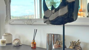 A room with a view of a cityscape through a window, a decorative mirror, a small table with various items including a candle, a shell, a bottle with a label, and a vase with sticks.