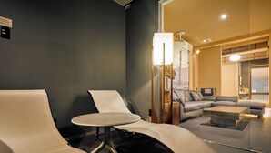 A modern hotel room with a dark blue accent wall, a round table, two lounge chairs, a sofa, and a glass partition.