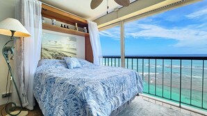 A bedroom with a bed, a nightstand, a lamp, a shelf with decorative items, and a large window with a view of the ocean.