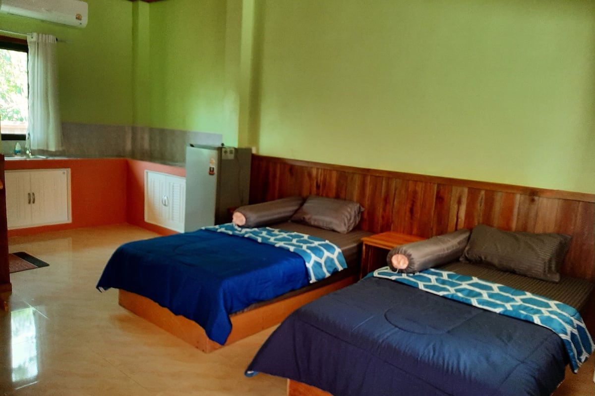 A room with two beds, a wooden headboard, a small table, and a kitchenette area.