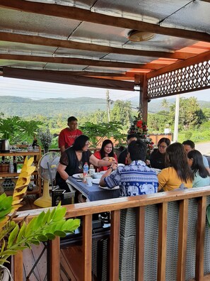 A group of people dining at an outdoor wooden table with a view of the hills. - Baan Berm Family Inn (Mae Rim)