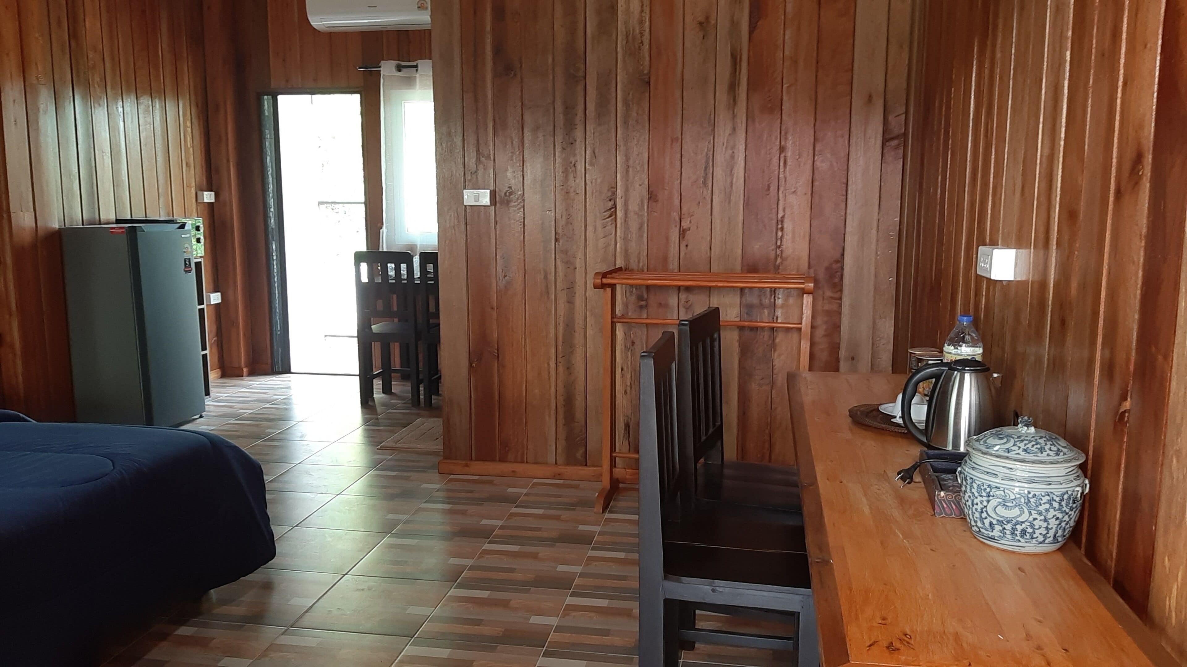A wooden-floored room with a bed, a chair, a table with a kettle, and a decorative pot.