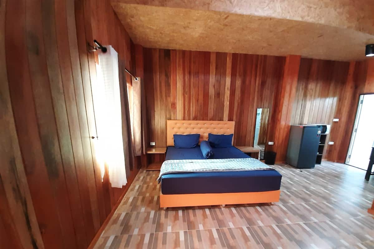 A bedroom with a wooden paneled wall, a large bed with blue bedding, a nightstand, and a refrigerator.