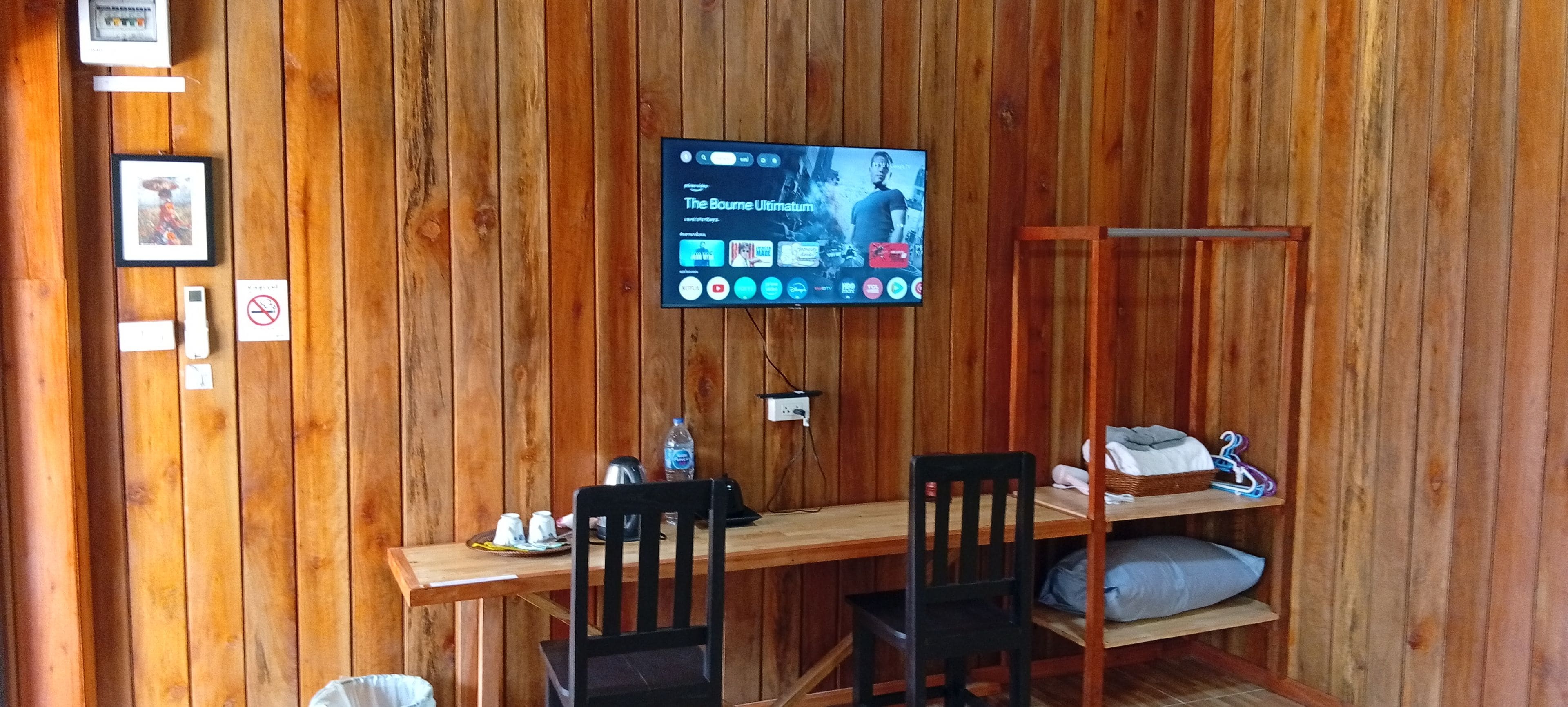Una habitación con paneles de madera, con un televisor montado en la pared mostrando una película. Hay una mesa de madera con sillas, un estante con objetos, y una pequeña estantería con un almohadón.
