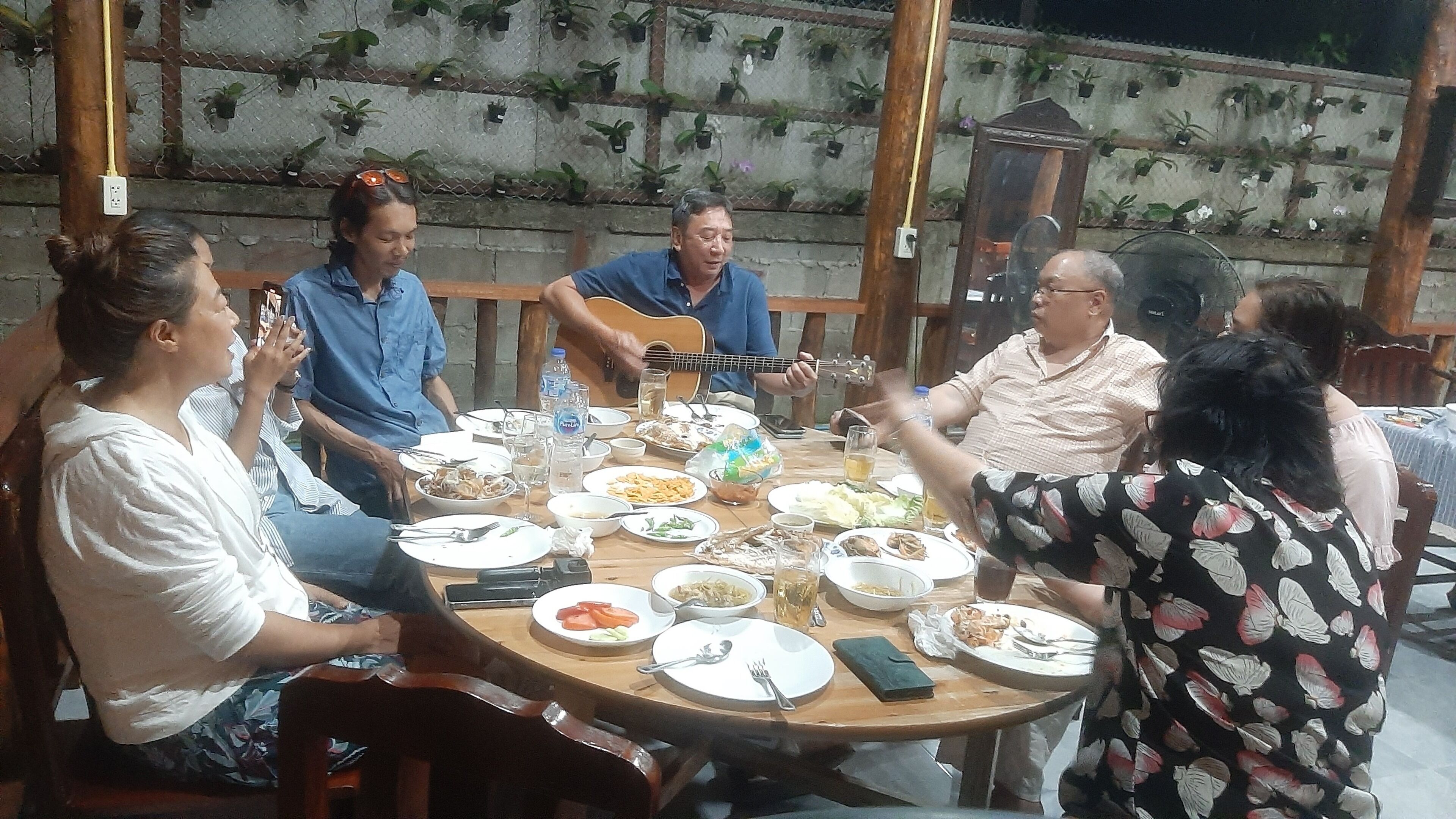 A group of people seated around a wooden table with various dishes and a guitar, in an outdoor setting with a concrete wall and potted plants.