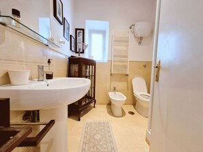 Ein Badezimmer mit einem großen, weißen Waschbecken, einem Bidet, einer Toilette, einem Glastischchen mit Toilettenartikeln und einem gemusterten Teppich auf dem Boden.