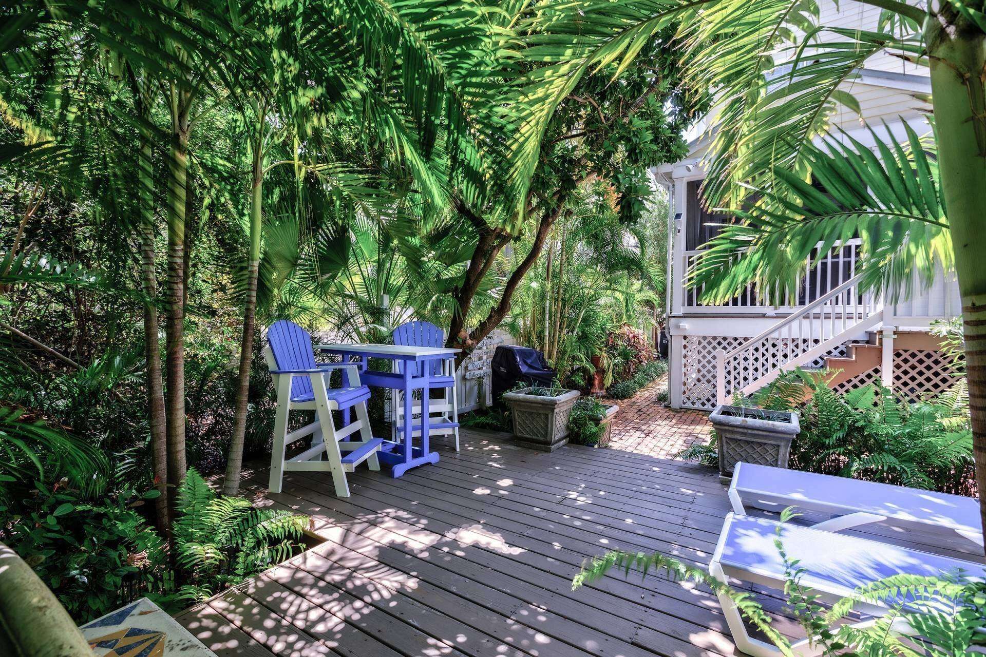 A wooden deck with a table and chairs, surrounded by lush greenery and a white fence.