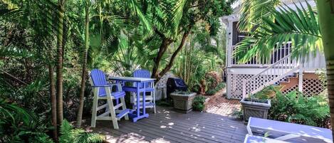A wooden deck with a table and chairs, surrounded by lush greenery and a white fence.