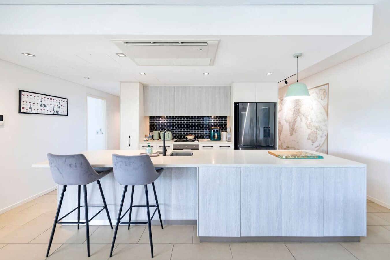 Une cuisine moderne dotée d’un grand îlot central, de deux tabourets de bar, d’un réfrigérateur en acier inoxydable et d’une carte du monde accrochée au mur.