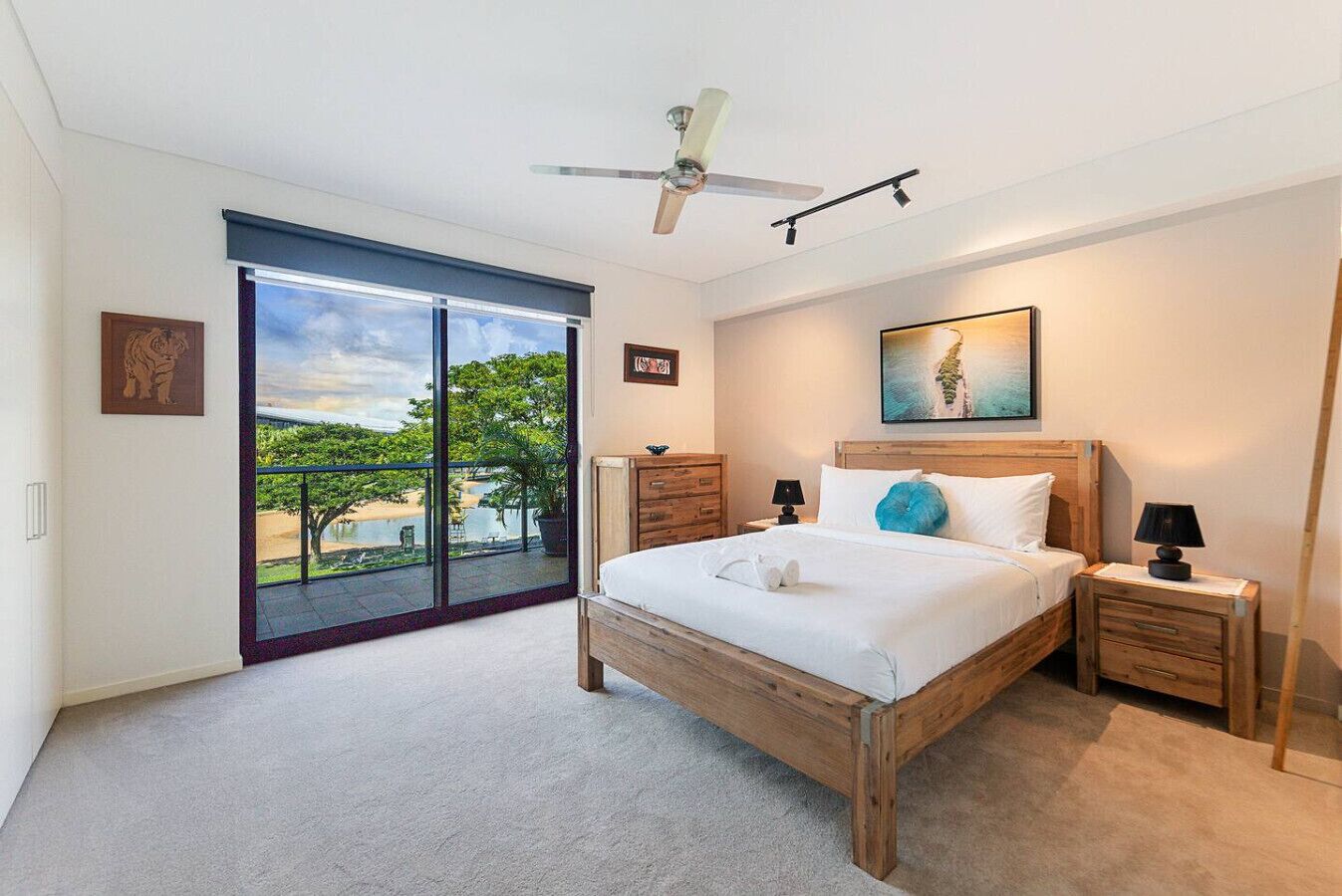 A bedroom with a large bed, a ceiling fan, a nightstand with a lamp, a wooden dresser, and a sliding glass door leading to a balcony with a view of trees and a body of water.