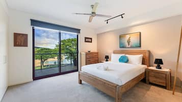 A bedroom with a large bed, a ceiling fan, a nightstand with a lamp, a wooden dresser, and a sliding glass door leading to a balcony with a view of trees and a body of water.