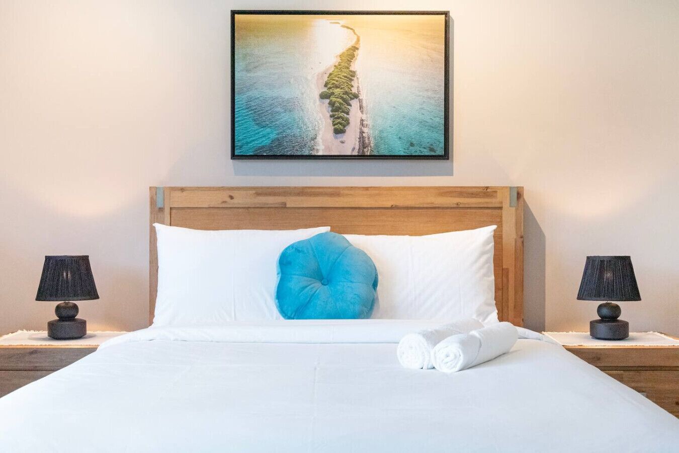 A hotel room with a wooden headboard, white bedding, a blue accent pillow, two bedside lamps, and a framed picture of a beach.