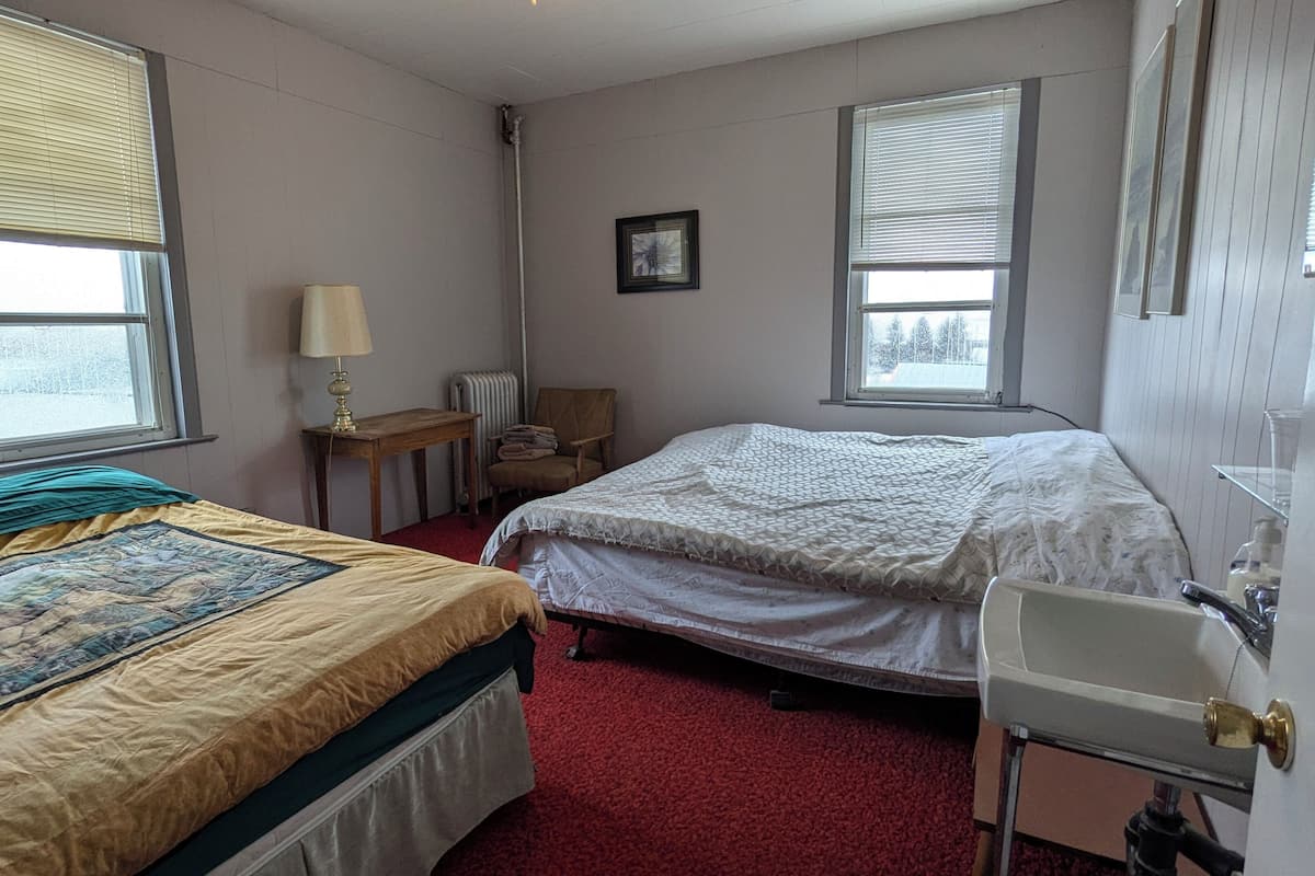 Un dormitorio con dos camas, una mesita de noche con lámpara, una silla y un ventanal.