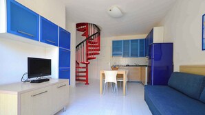 Ein kompakter Wohnraum mit Küchenzeile, einem Fernseher auf einem Schrank, einer roten Wendeltreppe und einem blauen Sofa.