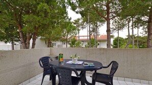 Ein runder Tisch mit vier Stühlen, einem Glas, einem Buch und einer kleinen Vase mit Blumen auf einem Balkon mit Fliesenboden und Bäumen im Hintergrund.