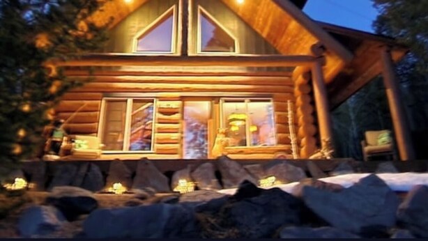 A log cabin with a prominent gabled roof, large windows, and a stone foundation.