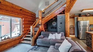 A log cabin interior with a wooden staircase, a gray sofa, and a kitchen area.