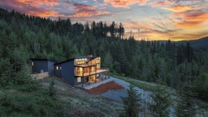Una casa moderna de dos pisos con un balcón grande, rodeada de un bosque frondoso y una vista panorámica al atardecer.