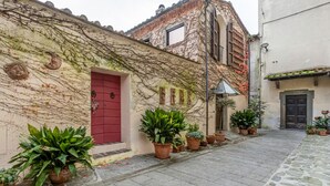A narrow cobblestone street with a building featuring a red door, potted plants, and ivy-covered walls. - GuestHost - San Rocco Apartment Borgo a Mozzano - Lucca (Borgo a Mozzano)