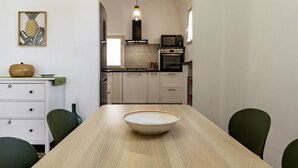 Ruokapöytä, jonka keskellä on kulho, taustalla keittiö kodinkoneineen, katosta roikkuva valaisin ja seinällä ananasta esittävä taideteos.