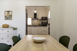 A dining table with a bowl in the center, a kitchen in the background with appliances, a hanging light fixture, and a pineapple artwork on the wall.