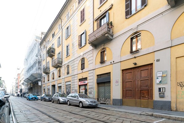 Una calle con coches estacionados y edificios con balcones y grandes portales.