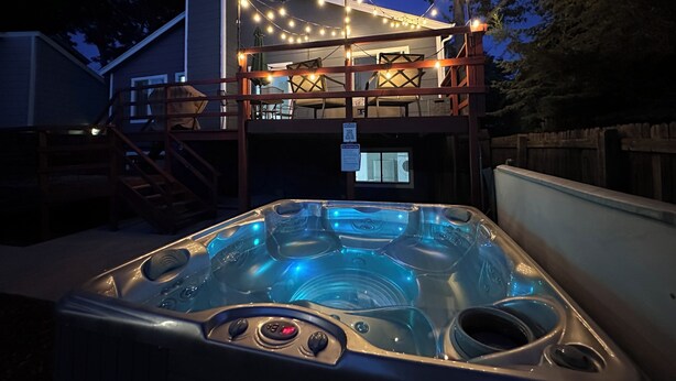 Een hottub met een houten terras en lichtjes erboven, omringd door een houten hek en bomen.