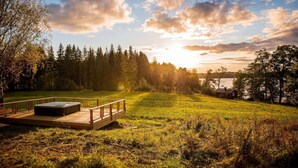 Una terraza de madera con un jacuzzi, rodeada por un campo cubierto de césped y un bosque.