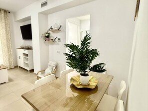 Un comedor con mesa de madera, sillas blancas, una planta en maceta y un televisor montado en la pared.
