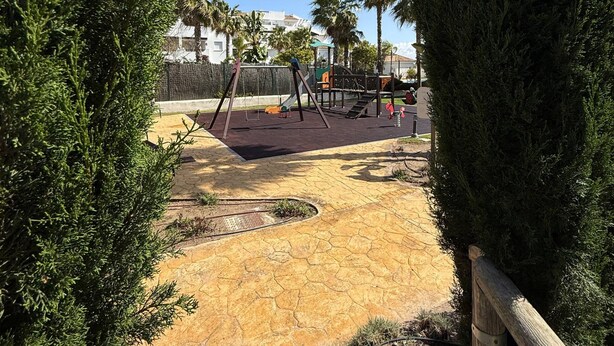 Un parque infantil con columpios y un tobogán, rodeado de árboles y un cielo despejado.