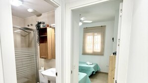 Un baño con ducha, inodoro, lavabo y un mueble pequeño. Hay una puerta que da a un dormitorio con dos camas y una ventana.