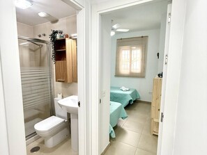 Un baño con ducha, inodoro, lavabo y un mueble pequeño. Hay una puerta que da a un dormitorio con dos camas y una ventana.