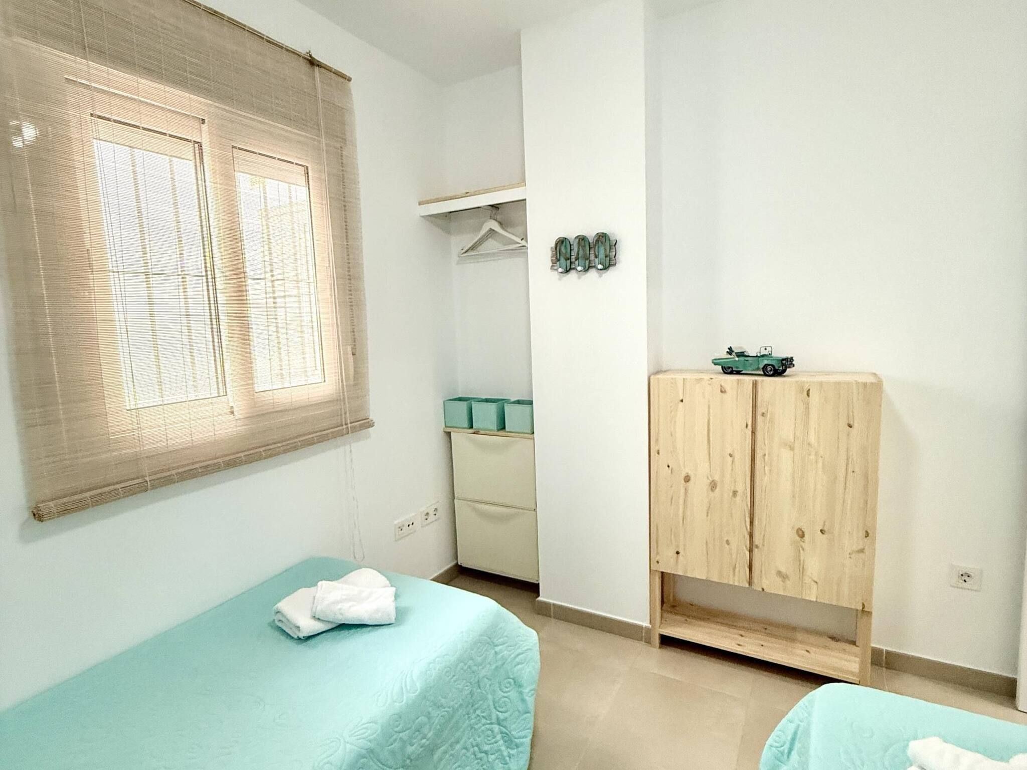 A bedroom with two beds, a wardrobe, a window with blinds, and a small shelf with decorative items.