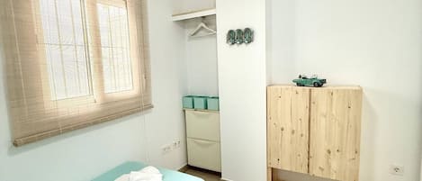A bedroom with two beds, a wardrobe, a window with blinds, and a small shelf with decorative items.