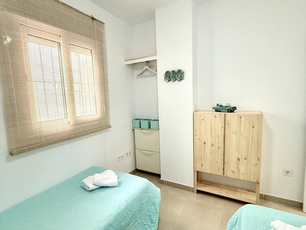 A bedroom with two beds, a wardrobe, a window with blinds, and a small shelf with decorative items.