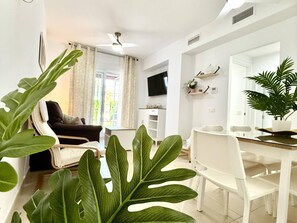 A modern living room with a sofa, dining table, chairs, and large green plants.