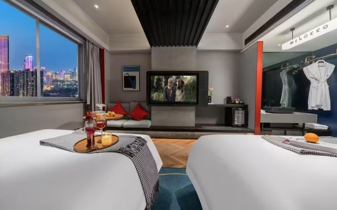 A modern hotel room with two beds, a TV, a city view, a robe rack, and a dining area with a table set for two.