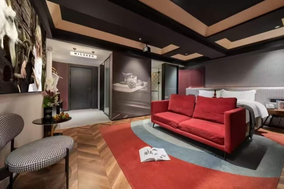 A modern hotel room with a red sofa, a wooden floor, a large mirror, and a wall-mounted TV.