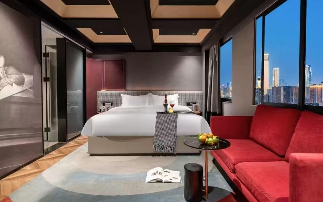 A modern hotel room with a large bed, a red sofa, a small table with fruit, and a city view.