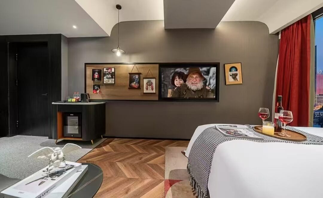 Ein modernes Hotelzimmer mit einem großen Bett, einem kleinen Schreibtisch und einem an der Wand montierten Fernseher, der einen Film zeigt.
