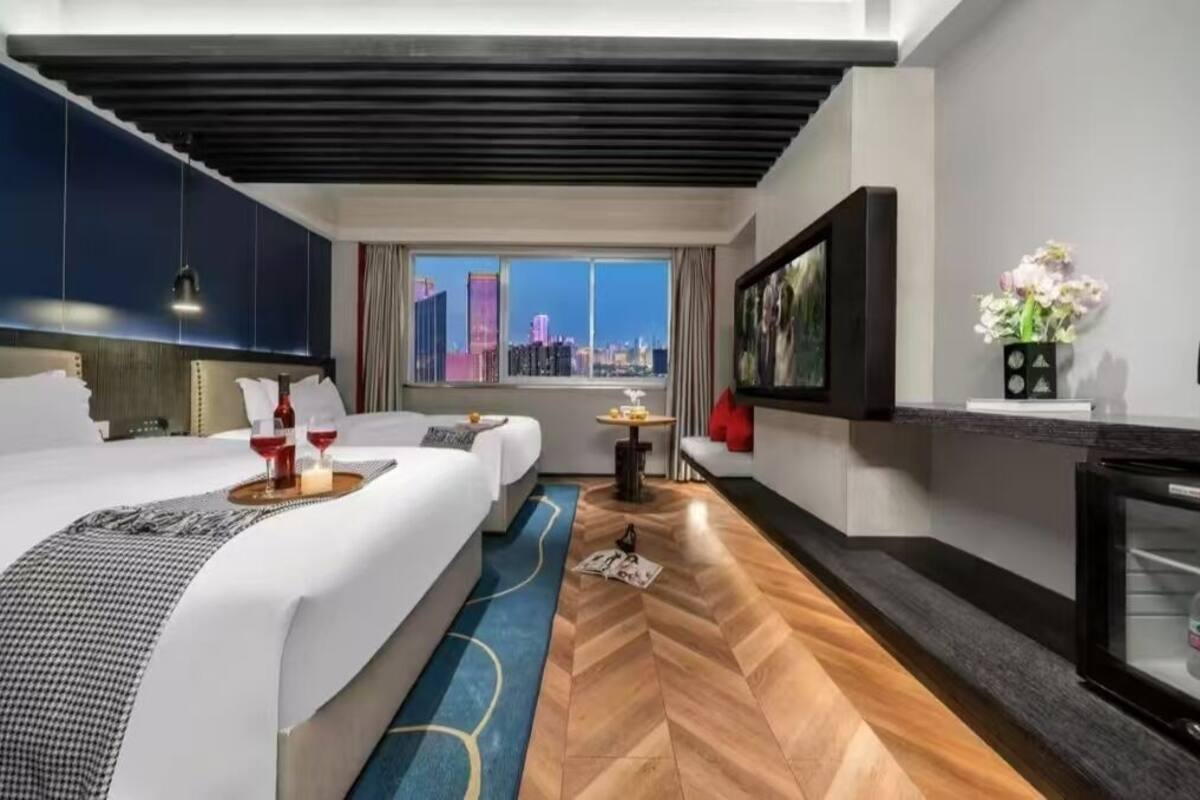 A modern hotel room with a large bed, a city view, a flat-screen TV, and a small table with a vase of flowers.