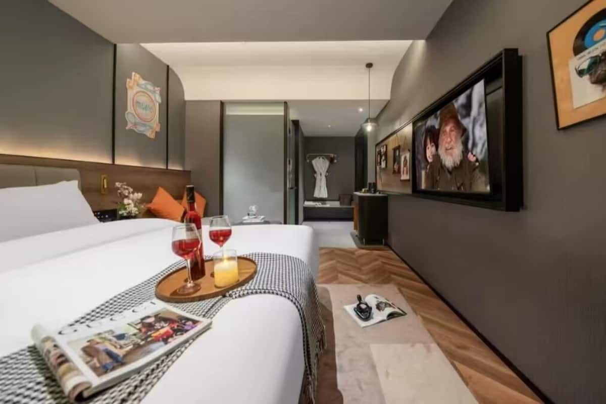 A modern hotel room with a neatly made bed, a side table with a magazine and wine glasses, and a large TV screen on the wall.