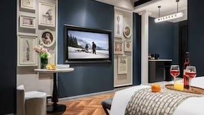 A modern hotel room with a large flat-screen TV, a small round table with a vase of flowers, a grey ottoman, and a bed with a patterned throw. - Milegu Hotel (Changsha)