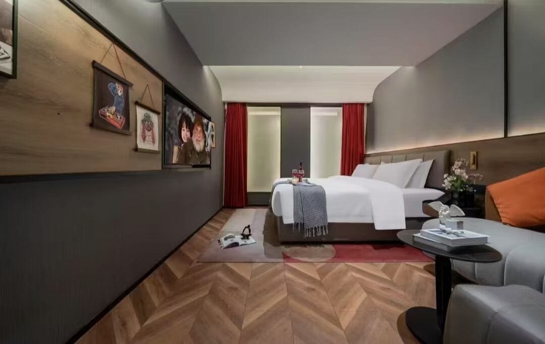 A modern hotel room with a large bed, a sofa, and a small table. There are framed pictures on the wall and a red curtain.