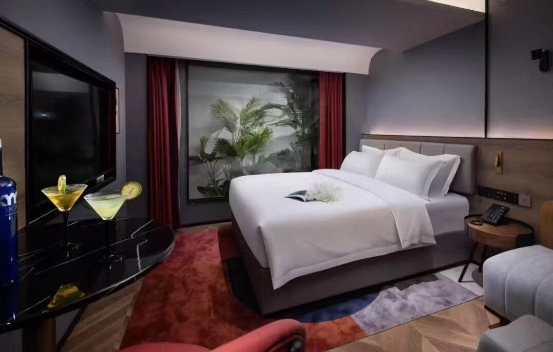 A modern hotel room with a large bed, a flat-screen TV, a minibar, and a window with a view of palm trees.